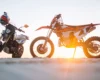An orange dirt bike and a black motorcycle parked on sandy ground at sunset