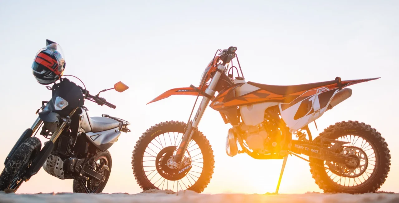 An orange dirt bike and a black motorcycle parked on sandy ground at sunset