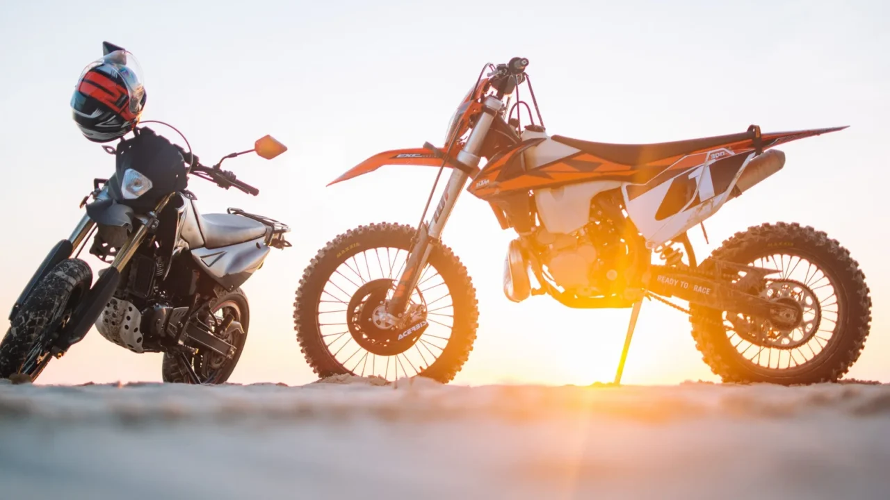 An orange dirt bike and a black motorcycle parked on sandy ground at sunset