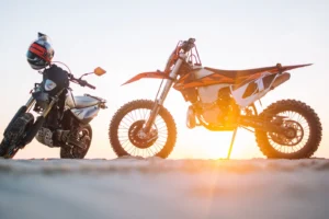 An orange dirt bike and a black motorcycle parked on sandy ground at sunset